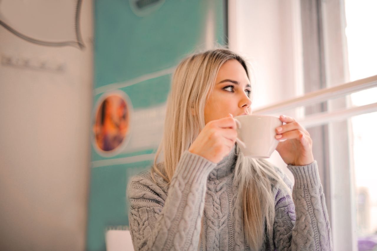 woman in gray sweater drinking from white ceramic mug while looking away